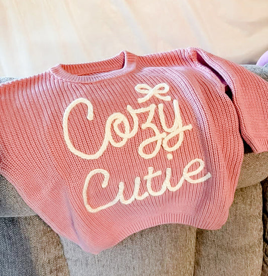 A pink ribbed knit sweater with 'Cozy Cutie' embroidered in cream script across the front, draped over a textured beige couch.